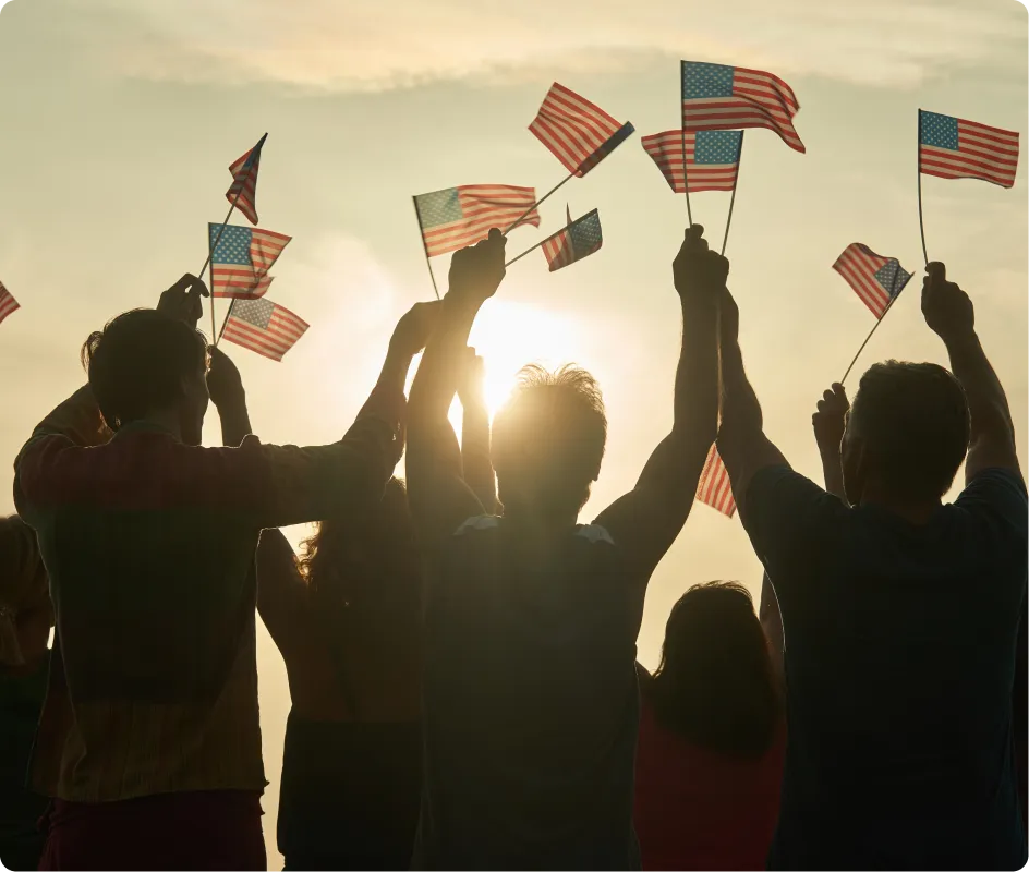 A group of people silhouetted against the sunset holds up small American flags, celebrating together outdoors.