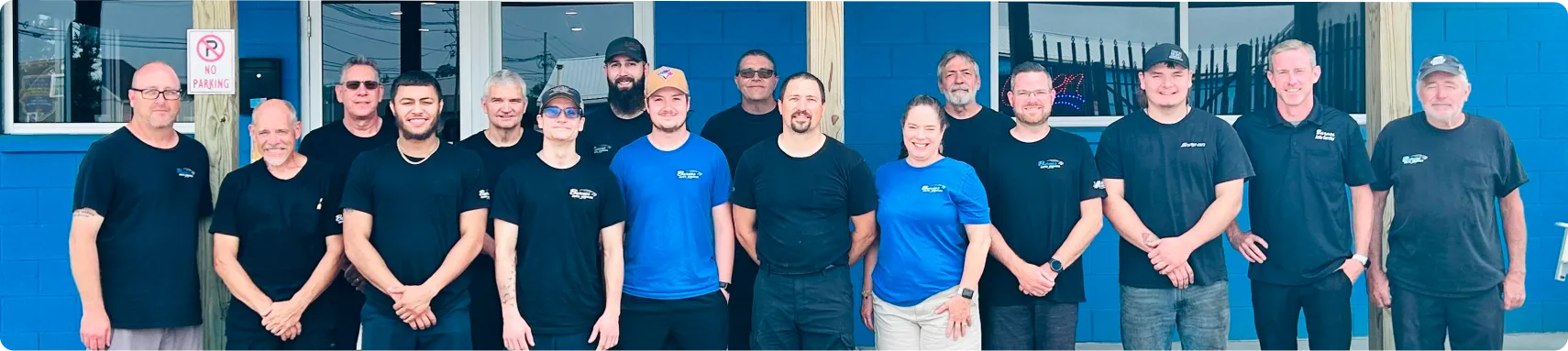 About Barnes Auto Service In Louisville, KY. A group of 17 people, mostly men and one woman, stand in front of a blue building, posing for a group photo. Most are wearing dark T-shirts, some with printed logos, and they are smiling at the camera.
