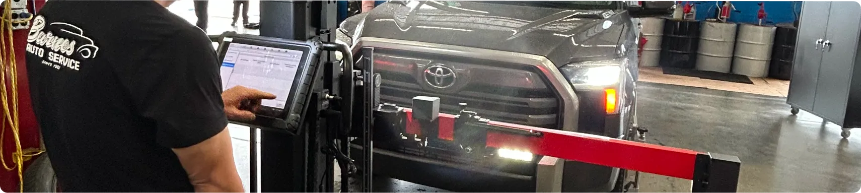 Diagnostics & ADAS Services In Louisville, KY At Barnes Auto Service. A mechanic uses a digital diagnostic tool to inspect a black Toyota pickup truck inside an auto service garage. The truck is positioned in front of alignment equipment.