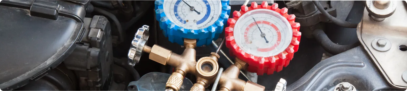Heat Repair, AC Repair In Louisville, KY At Barnes Auto Service. Close-up of a dual manifold gauge set with blue and red dials, used for measuring pressure in an automotive air conditioning system, placed on a car engine.