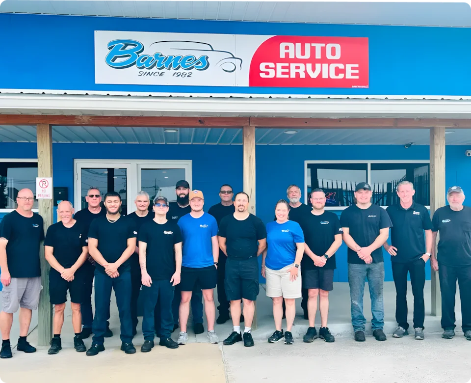 A group of fifteen people, wearing mostly black and blue outfits, stand smiling in front of a blue building with a sign that reads Barnes Auto Service. The building has a covered porch and large windows. Auto Repair In Louisville, KY At Barnes Auto Service