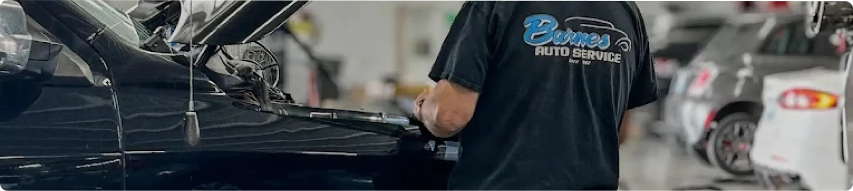 Diesel Repair In Louisville, KY At Barnes Auto Service. A mechanic wearing a “Benes Auto Service” shirt stands beside a black car with its hood open inside an auto repair shop, with other vehicles and equipment visible in the background.