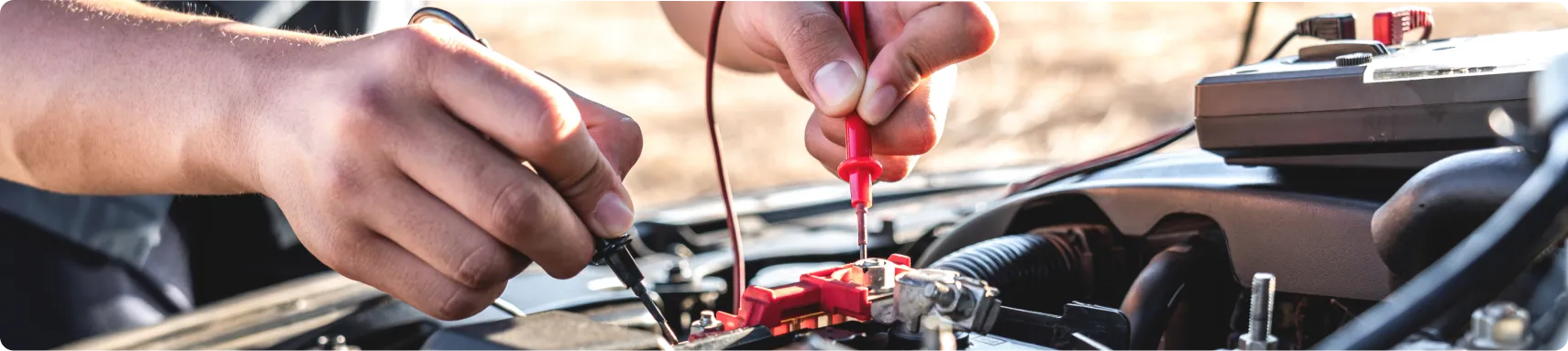 Auto Electrical Repair In Louisville, KY At Barnes Auto Service. A person uses a multimeter to check the voltage of a car battery under the hood, holding the probes to the battery terminals for testing.