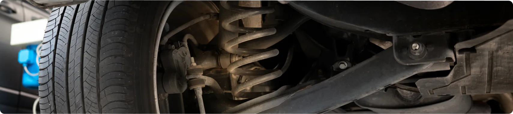 Suspension Repair In Louisville, KY At Barnes Auto Service. Close-up view of a car’s suspension system, showing the coil spring, shock absorber, and tire from underneath the vehicle in a garage or workshop setting.
