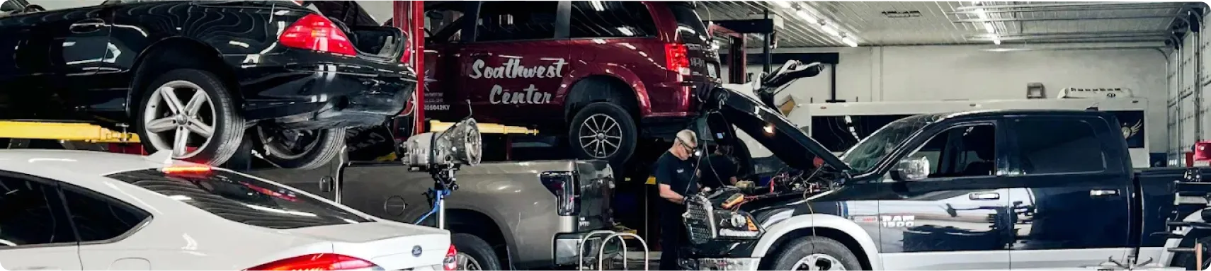 Vehicle We Service In Louisville, KY At Barnes Auto Service. A busy auto repair shop with several cars, SUVs, and a pickup truck being serviced by mechanics under bright overhead lights. Some vehicles are raised on lifts while others have open hoods.