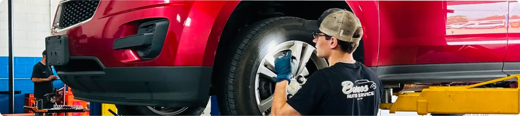 Vehicle Inspections In Louisville, KY At Barnes Auto Service. A mechanic wearing a cap and gloves inspects or repairs the front left wheel of a red car raised on a yellow hydraulic lift inside an auto repair shop.